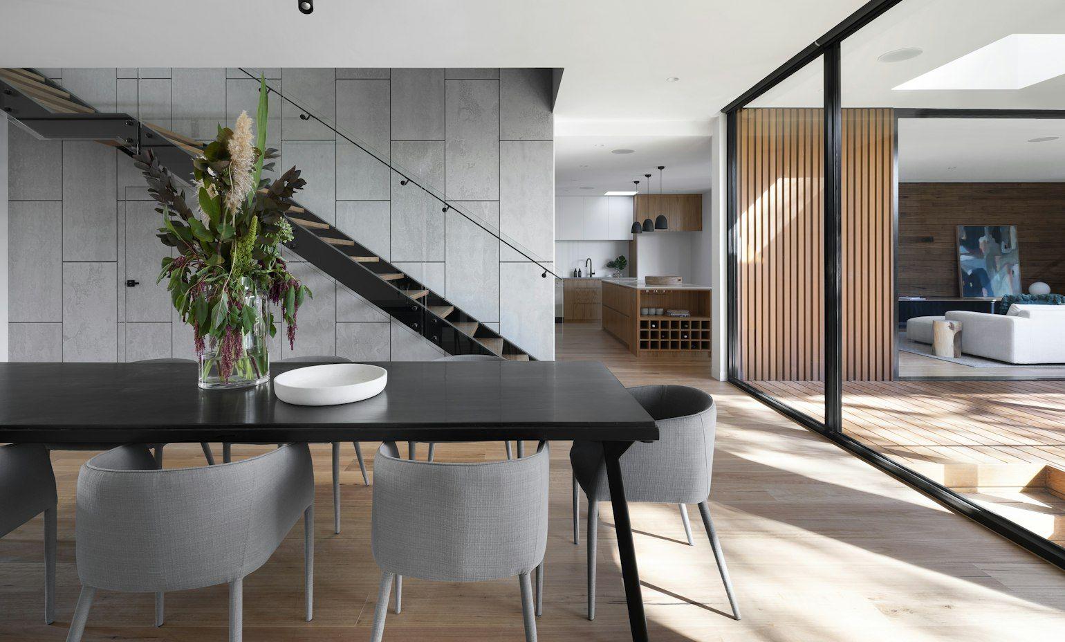 Dining area with a black table and grey chairs beside a floating staircase and timber-slat screen